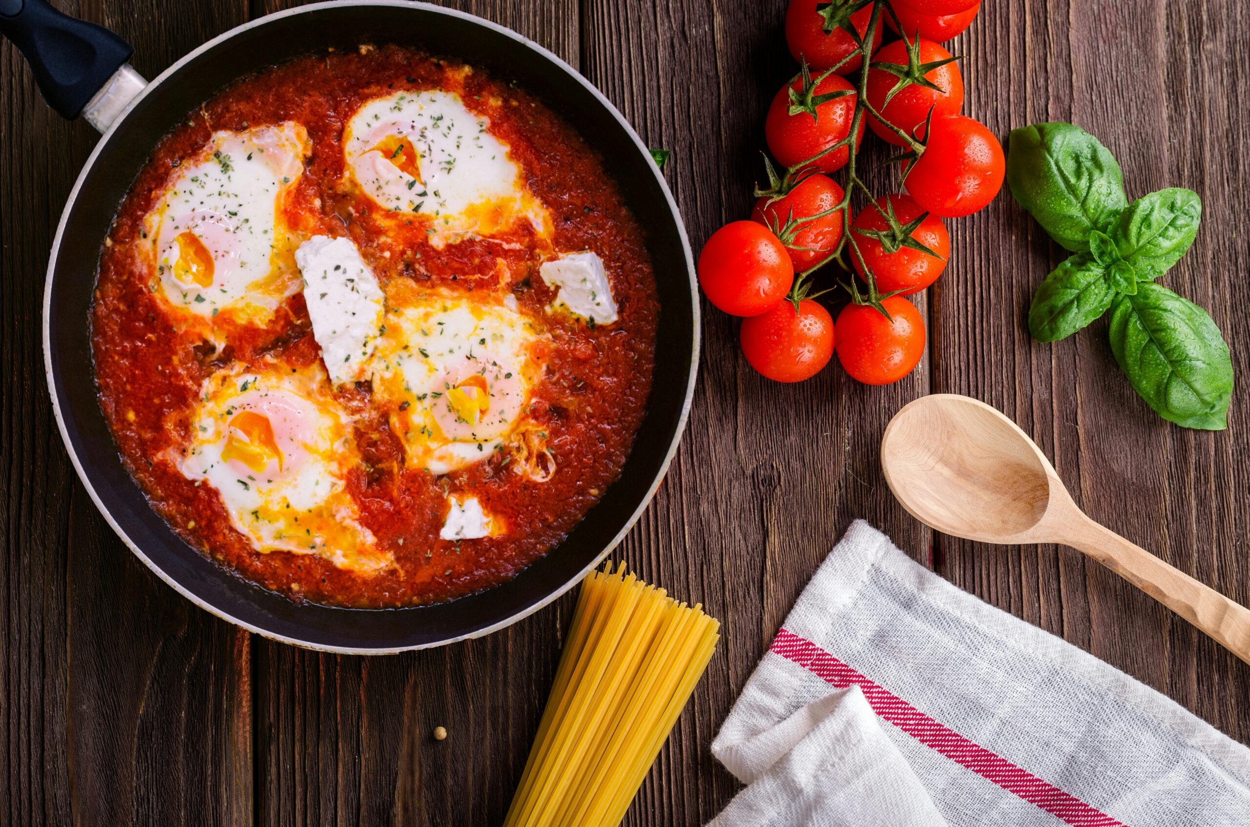 Overhead view of shakshuka in a black skillet on a rustic wooden table, featuring poached eggs in spicy tomato sauce, garnished with fresh herbs. Surrounded by cherry tomatoes on the vine, fresh basil leaves, dry spaghetti, a wooden spoon, and a beige linen napkin — perfect for a Mediterranean brunch or healthy breakfast recipe.